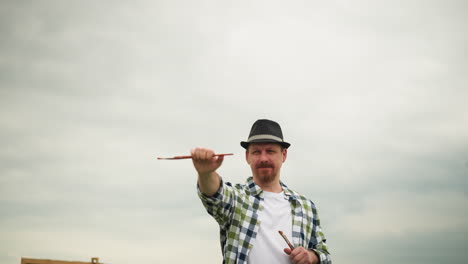 a woman in the foreground, slightly blurred, holds her hat while seated. in the background, an artist wearing a checked shirt and hat holds a paintbrush, pointing it forward to determine an angle