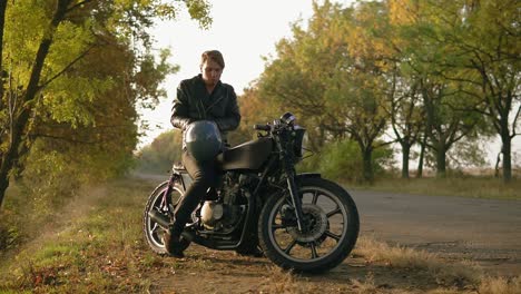 a young man in black leather jacket comes up to his motorcycle, sits there and puts a black helmet and sunglasses before