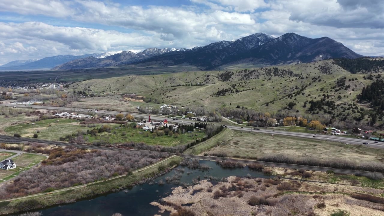 Una marisma de humedal refleja el pequeño pueblo agrupado de Bozeman a lo largo de una concurrida autopista; montañas distantes cubiertas de nieve se elevan más allá de colinas ondulantes cubiertas de maleza dorada de otoño. Toma aérea de dron de gran angular