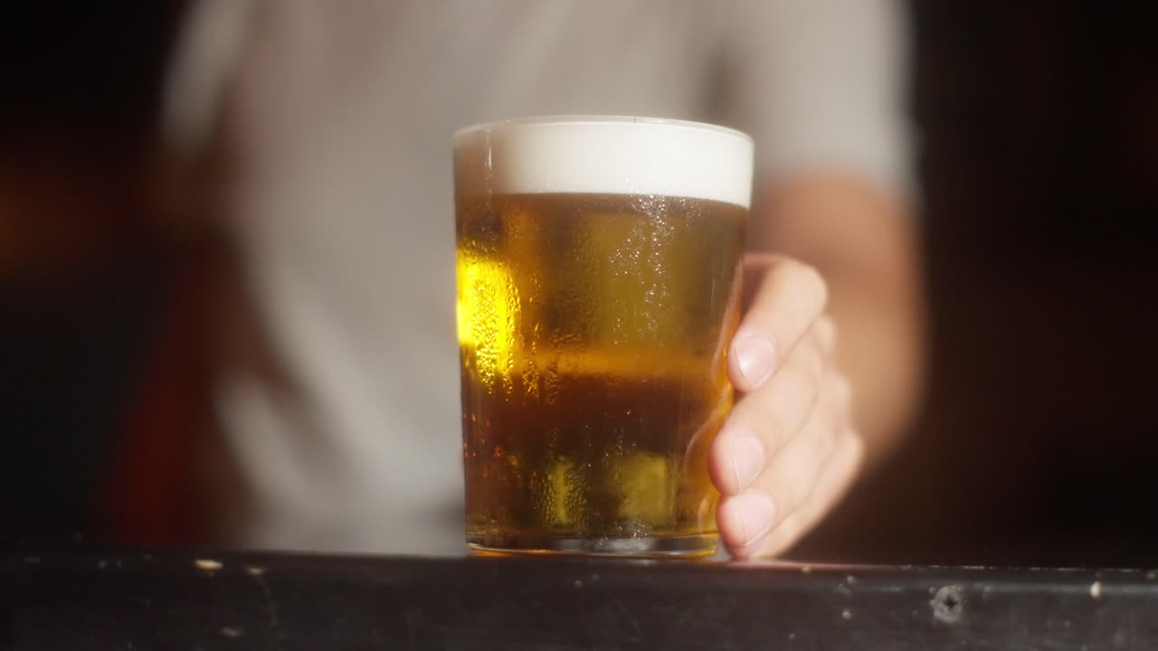 A beer glass comes into focus as a man in the background adjusts camera, starts out blurry