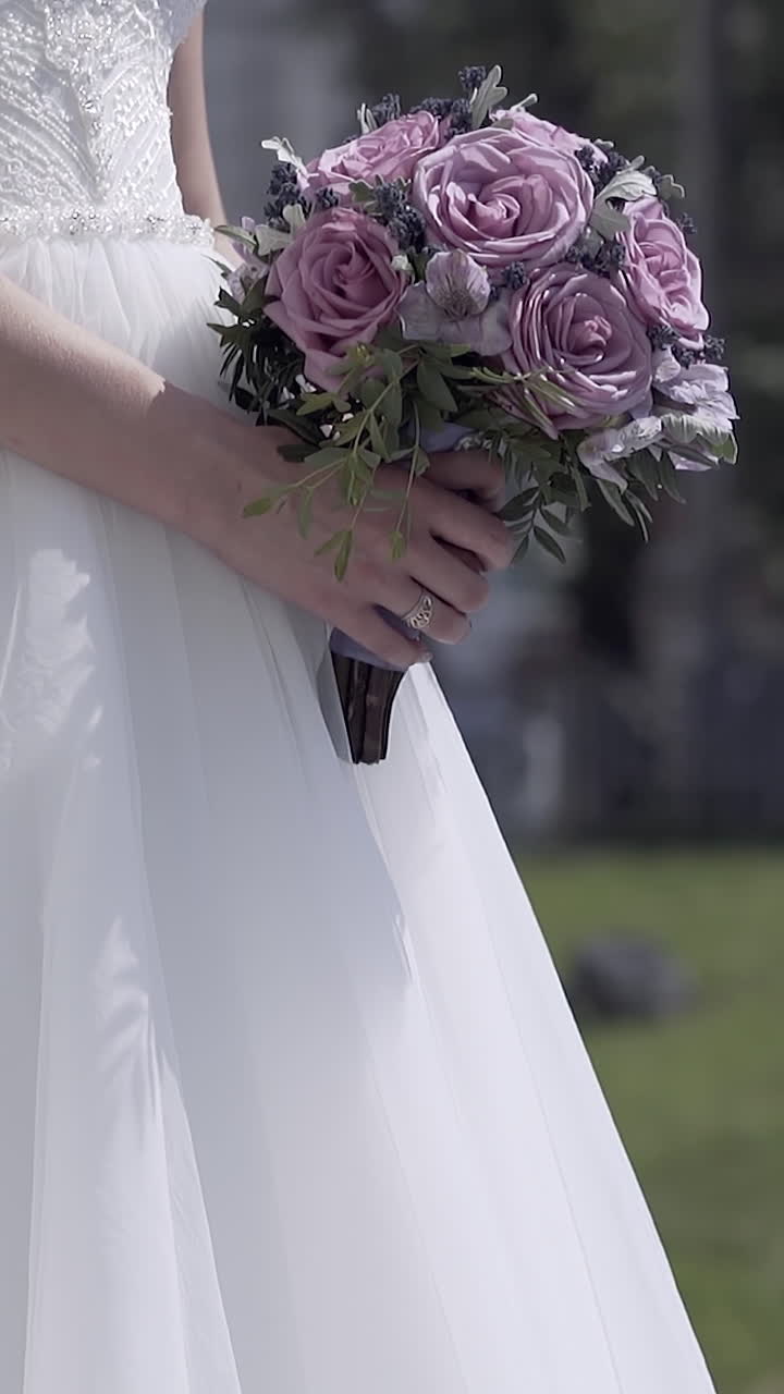 slow motion bride figure in white wedding dress with wonderful purple bouquet in hands against green grass