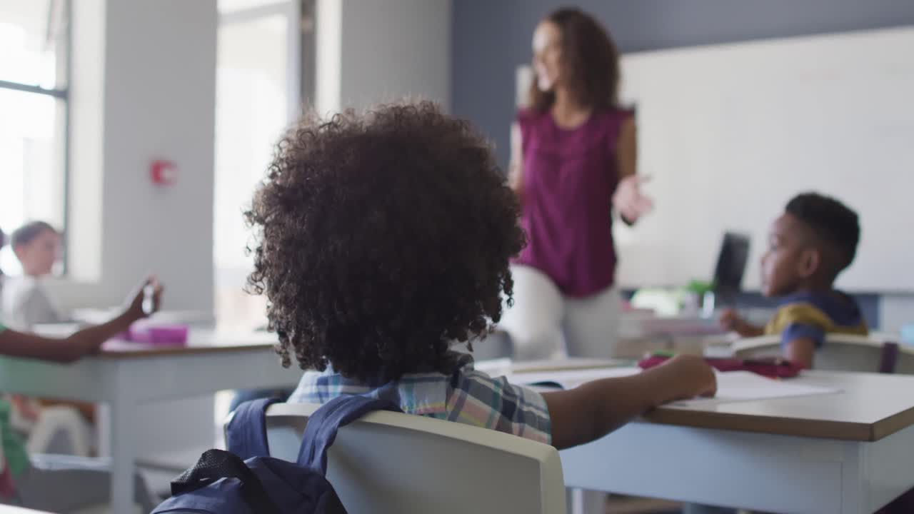 Video of back view of biracial boy studying in classroom with diverse pupils and teacher