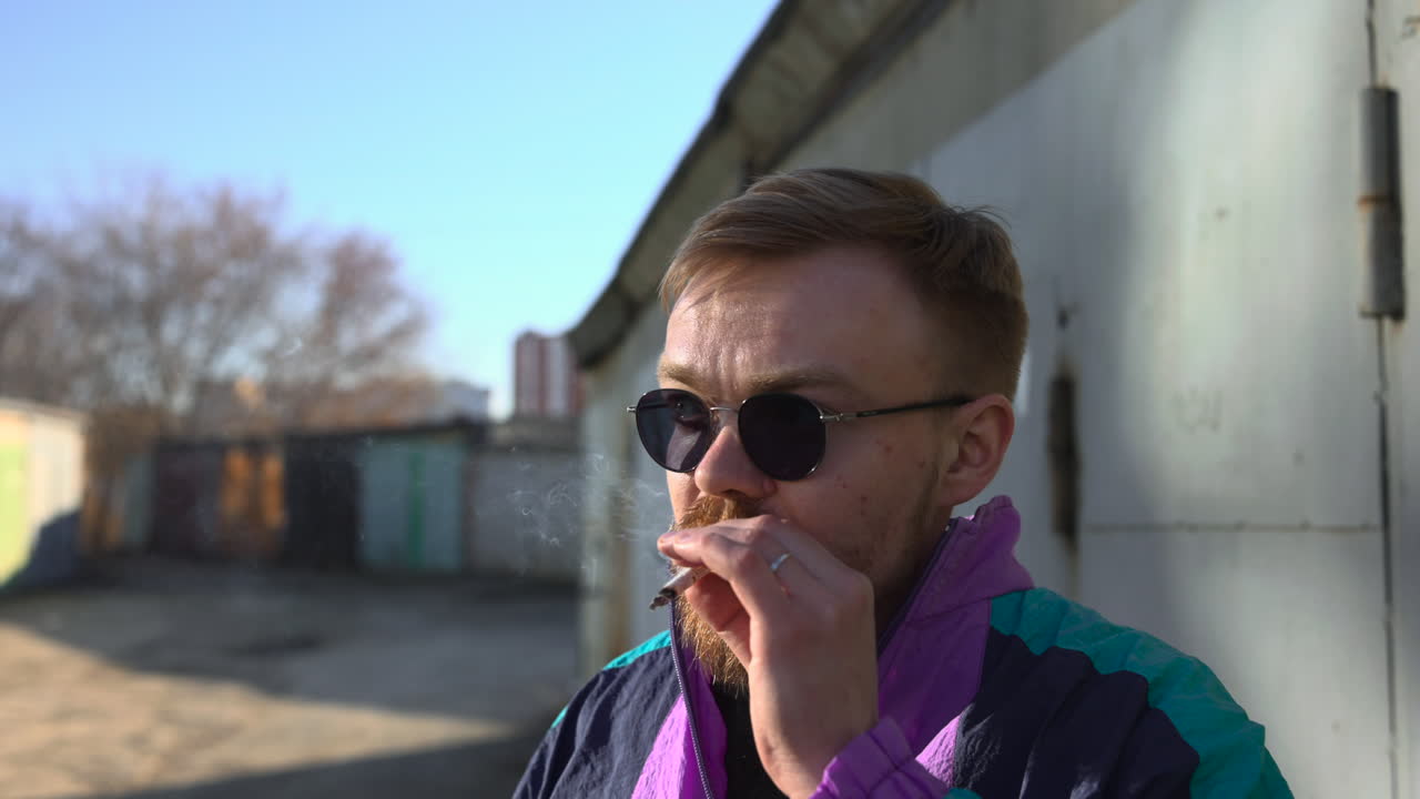 Uomo che ispeziona una sigaretta vicino a un muro di garage, tiene un filtro bianco, giacca a vento retrò, espressione pensierosa.