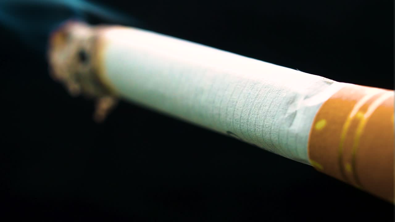 tailor made cigarette static on black backdrop burning from ne end white paper ashes falling down while smokes of blue colour are realised in the air yellow filter with golden marks and stripes sad