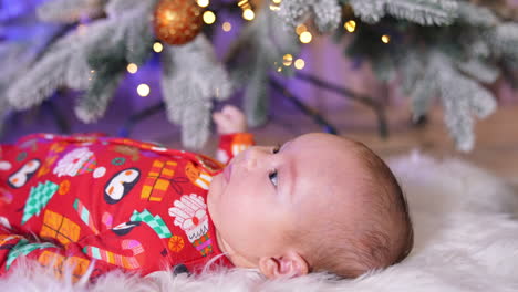 lovely little child in red clothes lies under christmas tree. adorable baby watches the shiny decoration dangling over him.