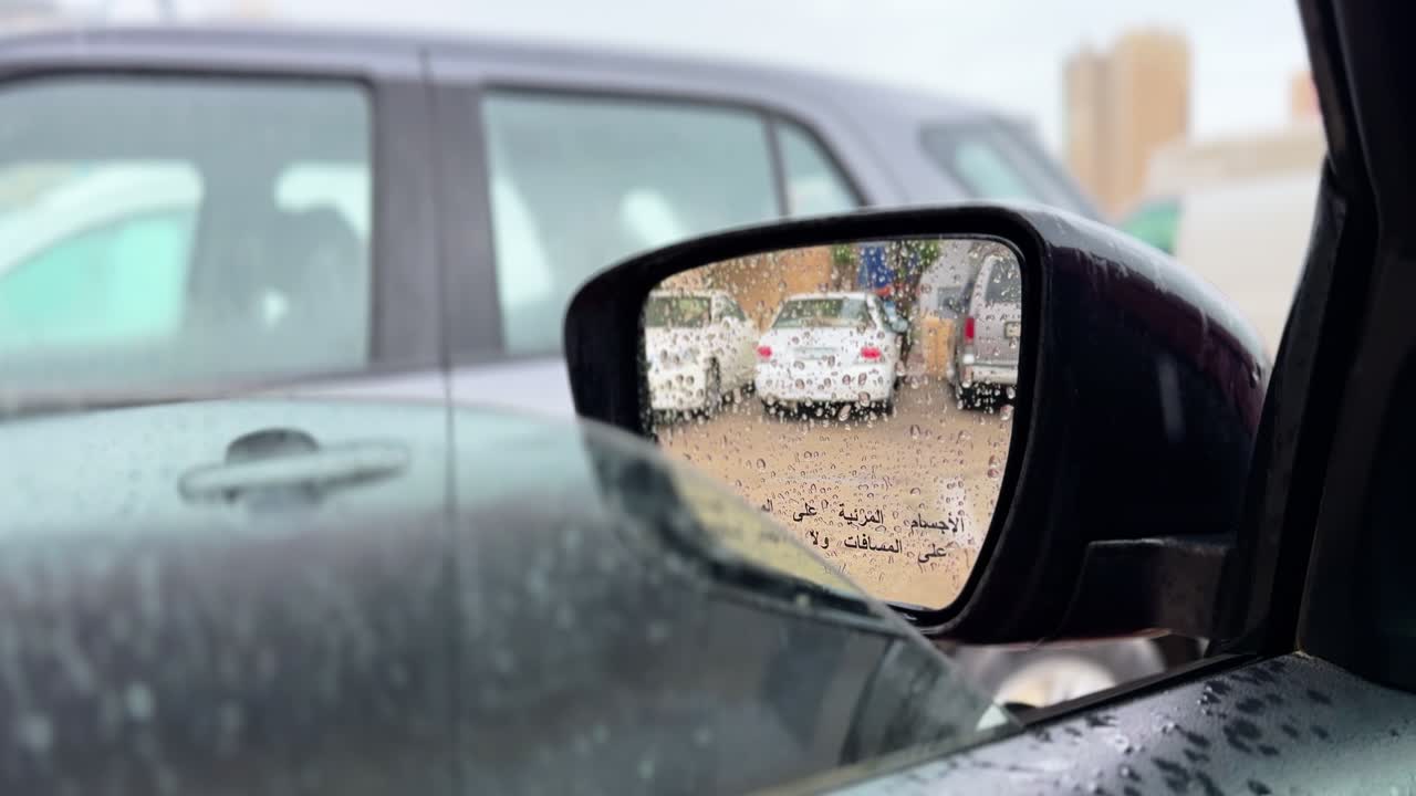 Dall'interno di un'auto, uno specchietto laterale coperto di pioggia riflette veicoli parcheggiati e il traffico su strada bagnata, catturando la guida urbana, la visibilità ridotta e le condizioni meteorologiche nuvolose.