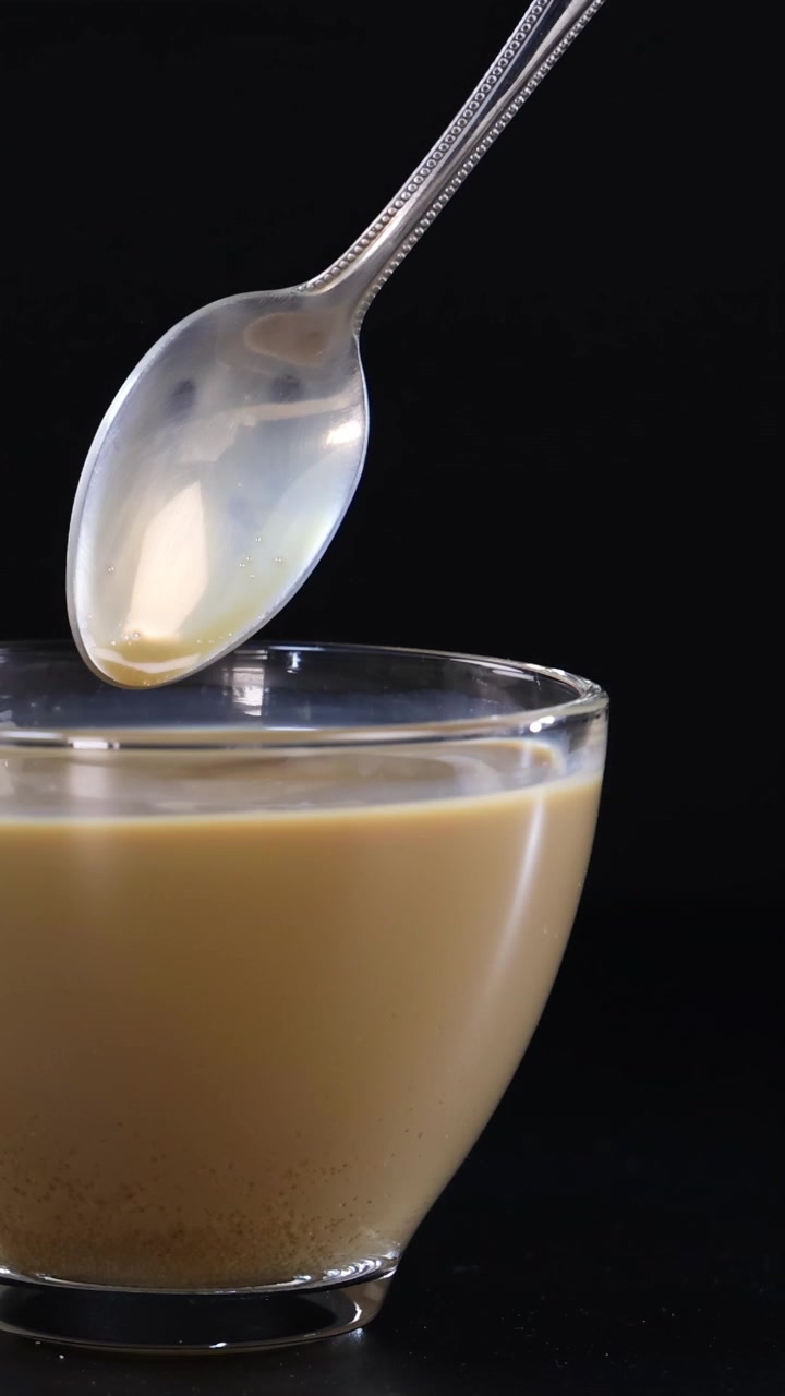 A hand stirs coffee in a glass cup using a spoon against a black background