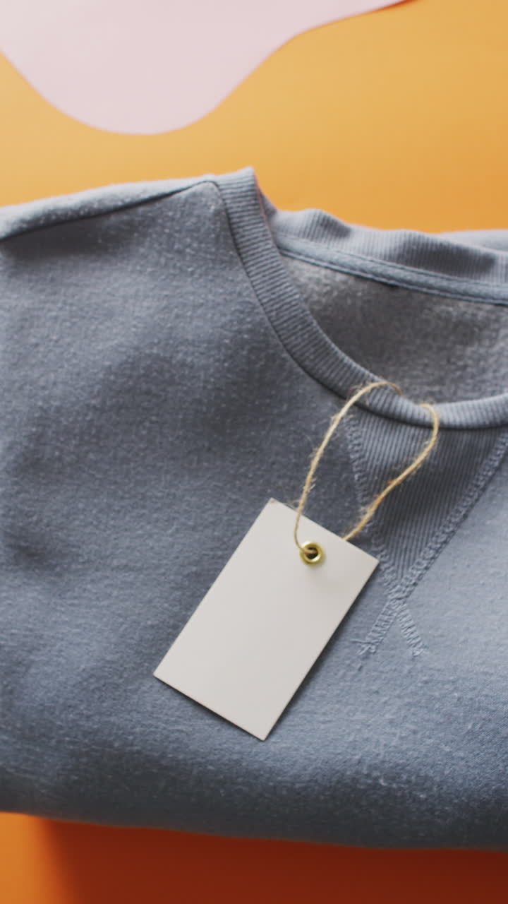 Vertical video of close up of blue folded t shirt with tag on orange background with copy space