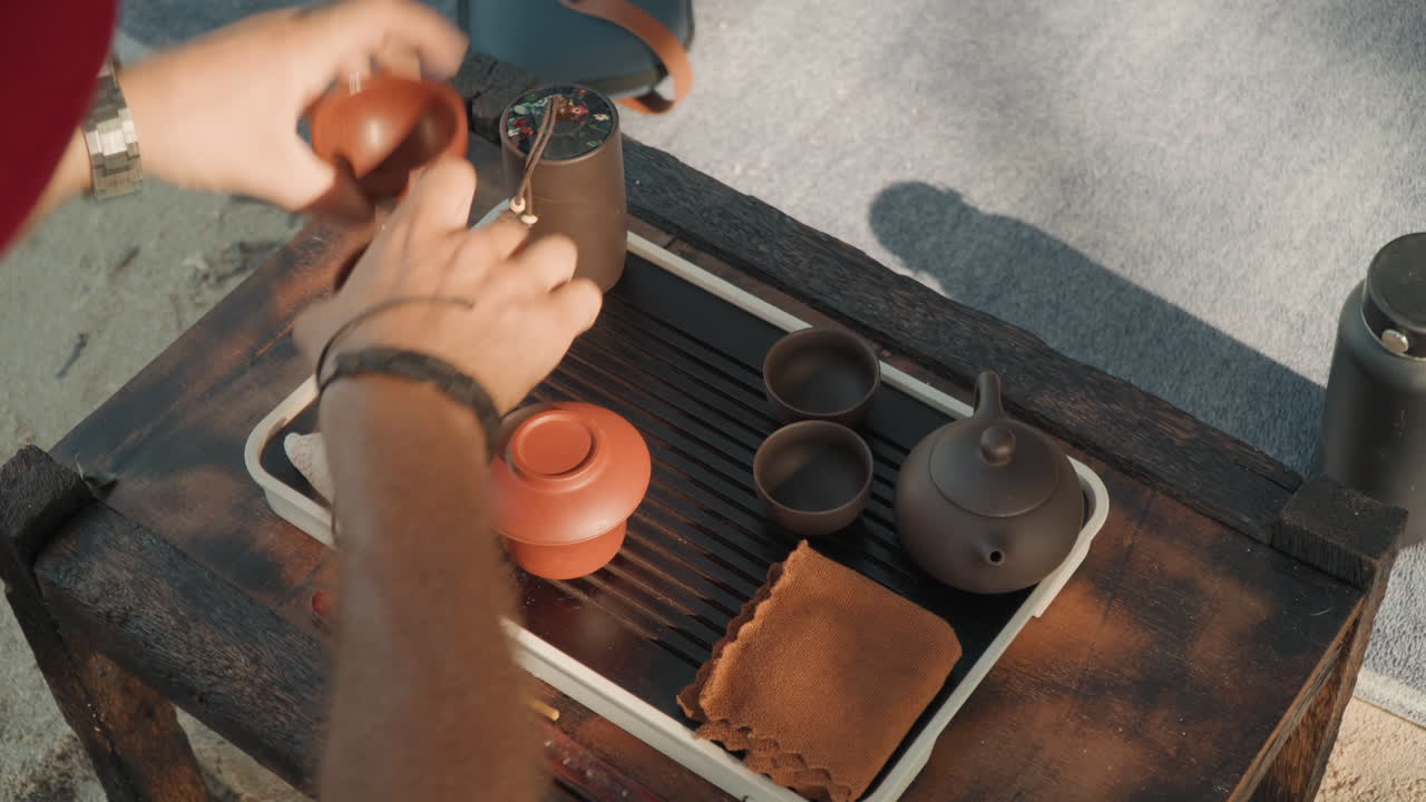 Hands arranging tea cups on tray at shoreline, clay cups handled with tongs, kettle and tea tools on rustic table, warm golden light, sandy surface, casual serving for small gathering, relaxed seaside