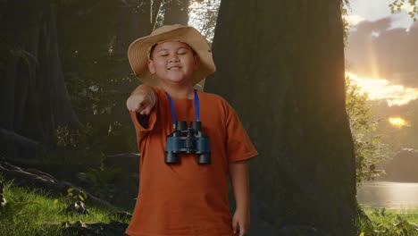 niño asiático con un sombrero y binoculares sonriendo y tocando su pecho luego señalándote mientras explora la naturaleza del bosque. niño investigador examina algo, turismo de viajes