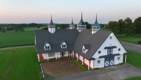 Aerial-view-of-Keeneland-Farms:-iconic-white-barn-with-pointed-turrets,-blue-trim-windows,-set-amid-lush-green-fields