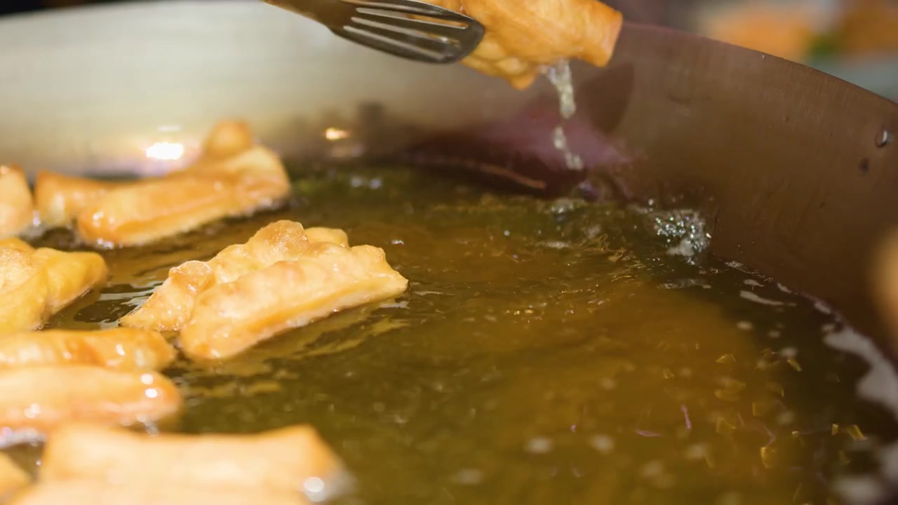 Nærbillede af gyldne dejstænger, der friteres i boblende olie, håndteret med metaltang.
