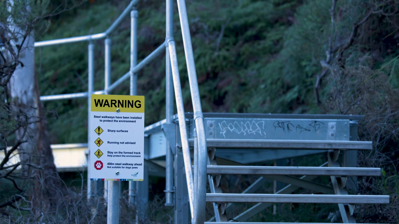 Primo piano di un segnale di avvertimento giallo su scale d'acciaio lungo un sentiero escursionistico a Wentworth Falls