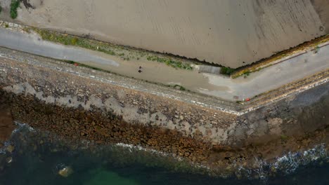 Bird's-eye-view-of-Spiddal-East-Pier,-with-waves-breaking-in-as-drone-follows-pedestrians