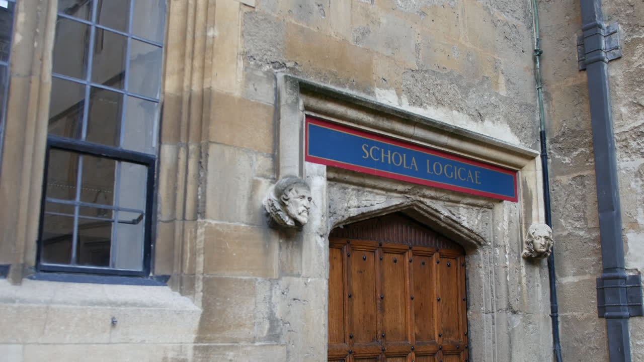 Ingresso con portale all'Università di Oxford con la scritta "Schola Logicae" che significa scuola di logica, caratterizzato da teste di pietra intagliate, pannelli di porte in legno e superfici architettoniche usurate.