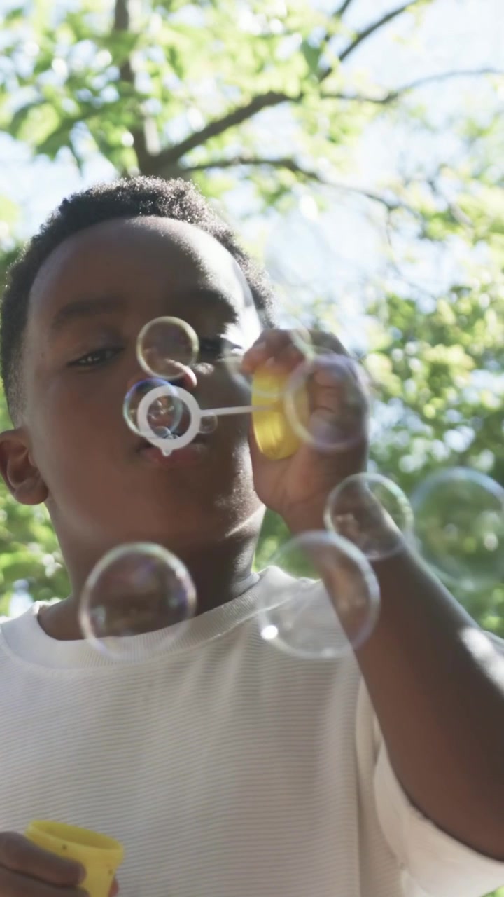 Vertical video: Lifting yellow wand, boy wearing white tee blowing bubbles in yard with sunlight