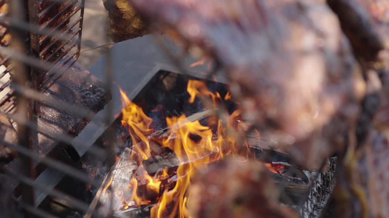 Flames dance under sizzling meat in a traditional Argentine asado, where iron racks and wood embers frame the intense cooking scene of open-fire grilling in a rustic outdoor setting In slow motion