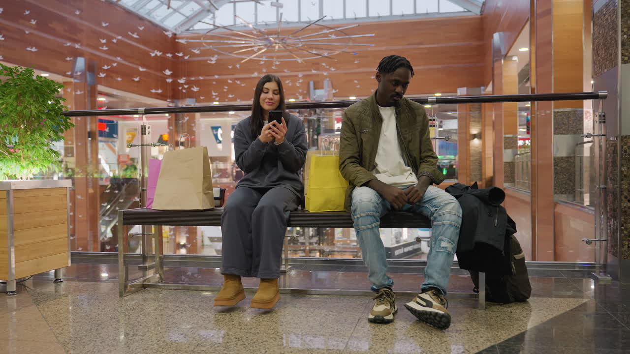 Unhappy young man sits beside cheerful woman using smartphone on mall bench with shopping bags, modern mall background, natural light from skylight and wooden panels
