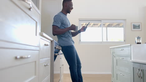 feliz hombre afroamericano bebiendo café y usando teléfono inteligente en una cocina soleada, en cámara lenta