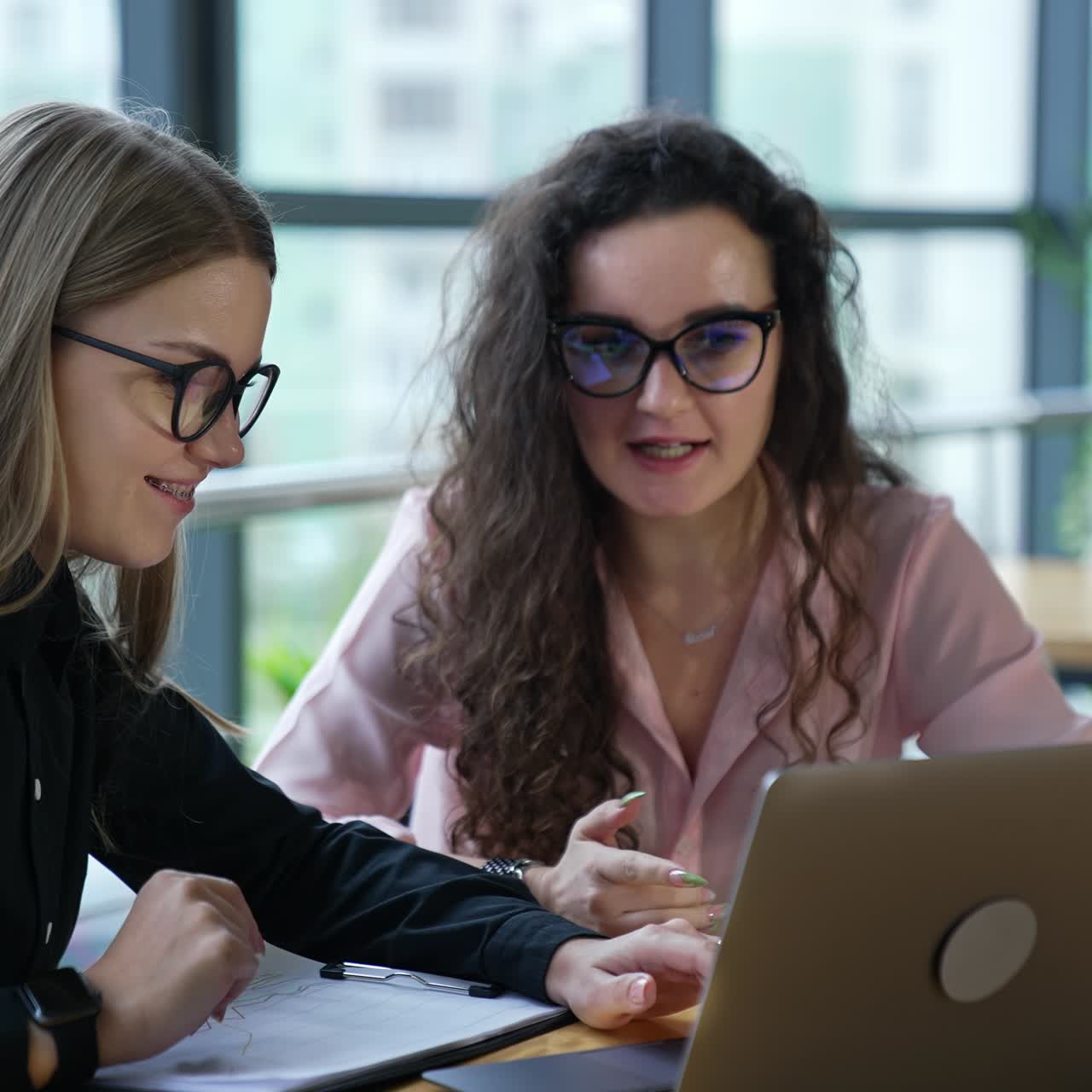 Women coworkers discuss working moments at the table. Female employees look at laptop and talk. Blurred office background