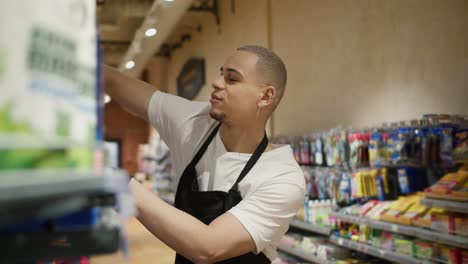 handsome worker in apron arranging shelves with products from the cart