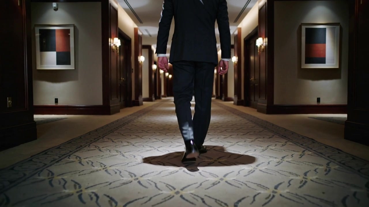 Ripresa in movimento che segue un uomo adulto mentre cammina verso un'insegna luminosa nel corridoio di un hotel, con abito elegante e moquette
