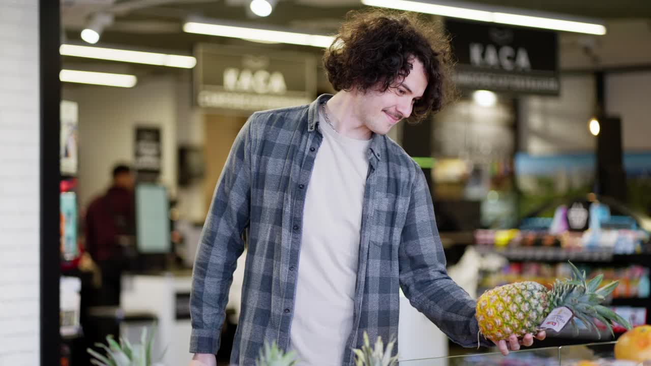 Happy brunette guy with curly hair in a plaid shirt examines products in the citrus department of pineapple and orange fruits in the supermarket