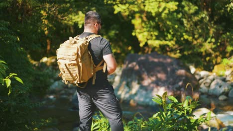 Young-male-hiker-looking-around-during-hike-in-jungle-by-a-river-surounded-by-boulders-and-trees-hike-in-wild-forest-hot-summer-day,-dolly-truck-shot