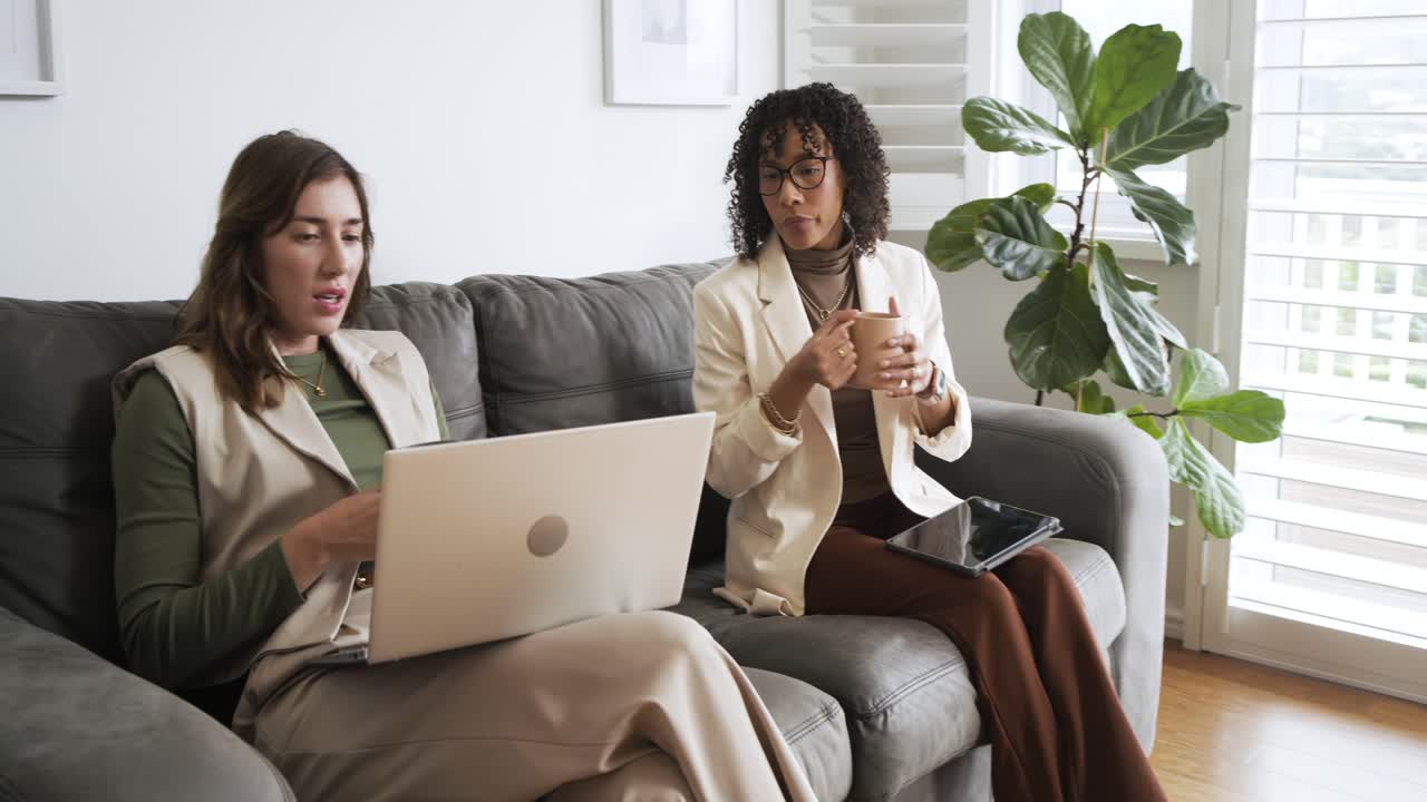 Collègues de travail diversifiés assis sur un canapé, sirotant des tasses, tapant sur une tablette, saisissant sur un ordinateur portable et discutant d'un projet.