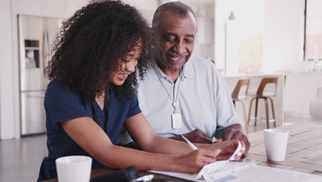 Smiling-female-healthcare-worker-filling-in-paperwork-with-senior-male-patient-during-a-home-visit