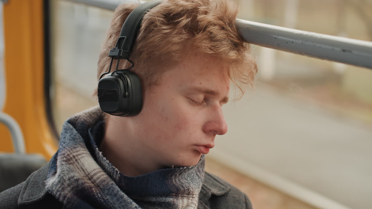 Primo piano di un uomo bianco, creatore, con le cuffie, di profilo su un tram in movimento con gli occhi chiusi. Sciarpa avvolta al collo, luce calda e soffusa, ispirazione pacifica e concentrazione silenziosa, produttore musicale.