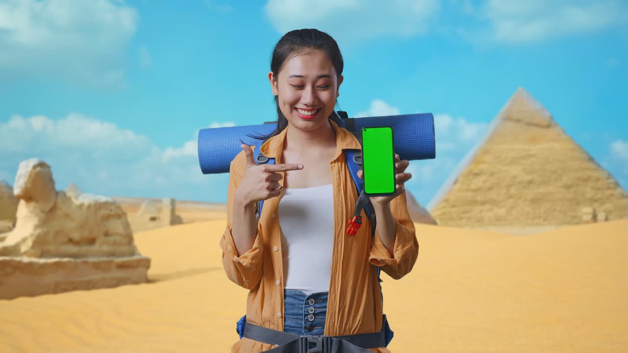 Asian Female Hiker With Mountaineering Backpack Smiling And Pointing To Mock Up Green Screen Smartphone While Traveling In Pyramid Of Giza