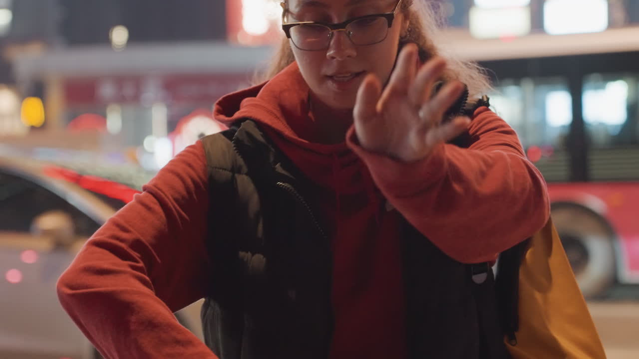 Young woman under neon signage night gesturing to passersby pointing toward transit stop expressive hand movements streetwear hoodie backpack candid urban interaction vibrant glow