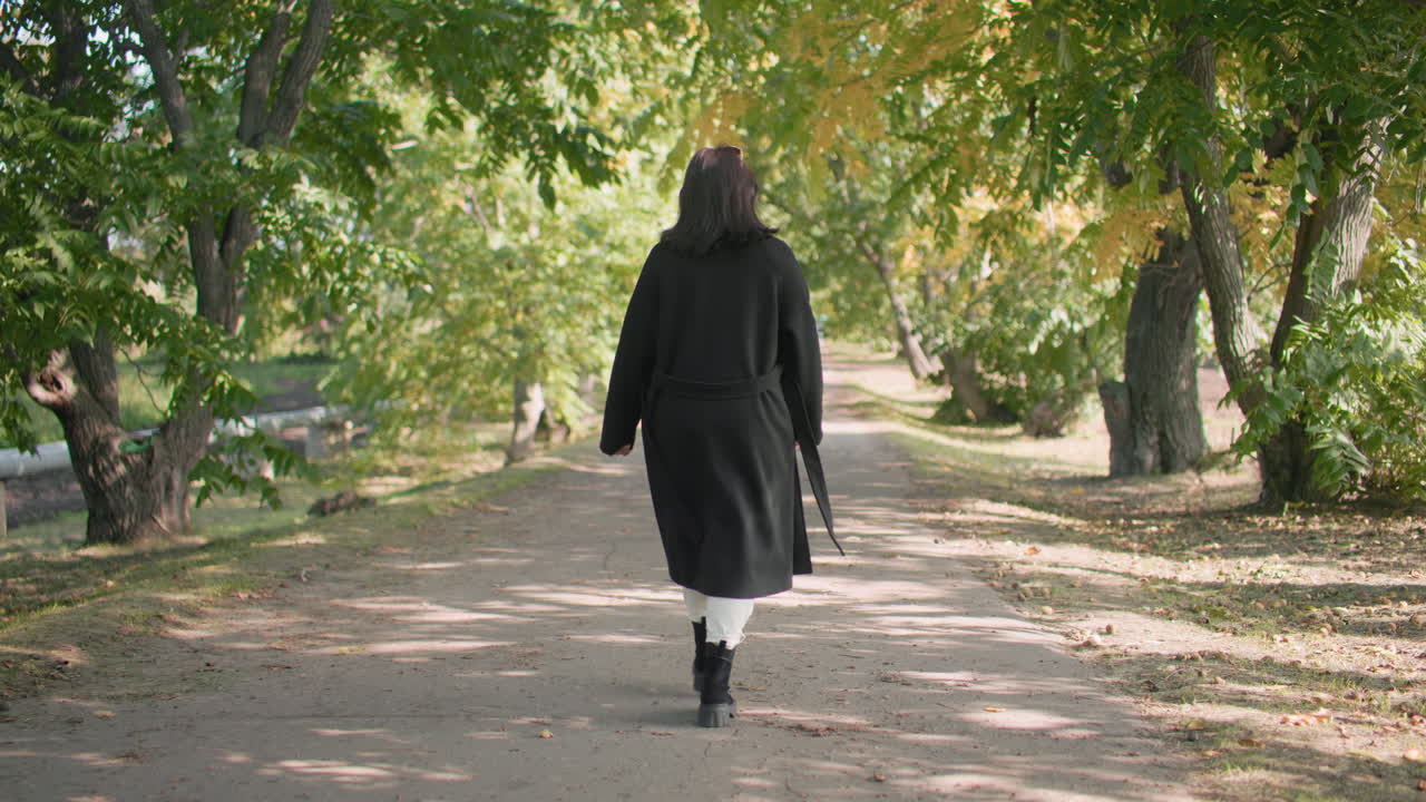 Back view of contemplative girl in long black coat walking along arboretum path surrounded by lush trees, autumn sunlight filtering through branches, enjoying peaceful moment
