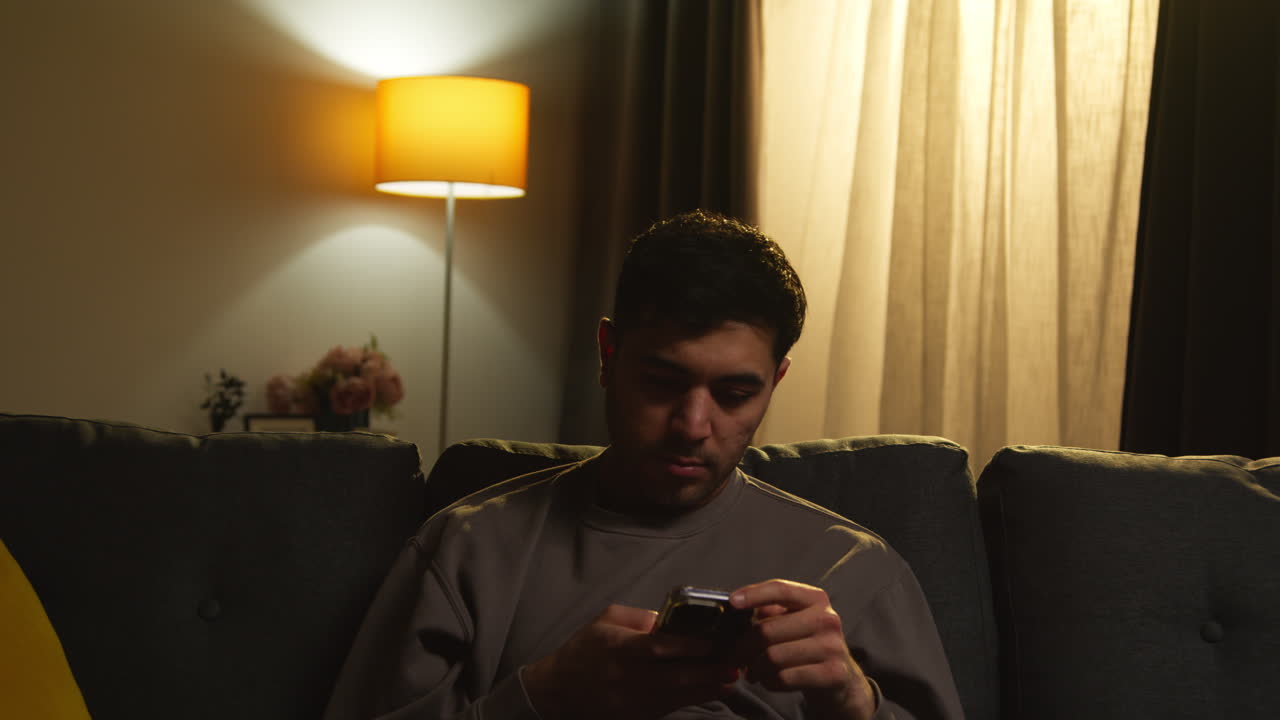 joven que pasa la noche en casa sentado en el sofá con el teléfono móvil explorando internet o las redes sociales 2