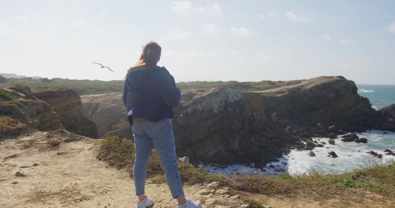 A wide slow-motion 4K shot of a person in a blue coat observing the dramatic Peniche coast, with the ocean crashing against the rocks in a windy, expansive landscape
