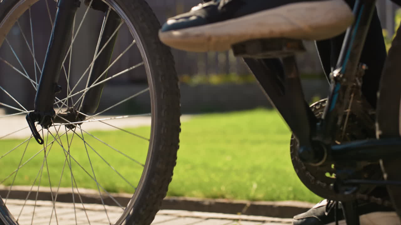 Forhjul, der kører langs en solbeskinnet kantsten med jævn pedal og roterende eger, sneaker, der trykker pedalen ned, skarpe detaljer på fælg og nav med manicured græsplæne og hegn som baggrund, livlig eftermiddagsrytme og fokuseret