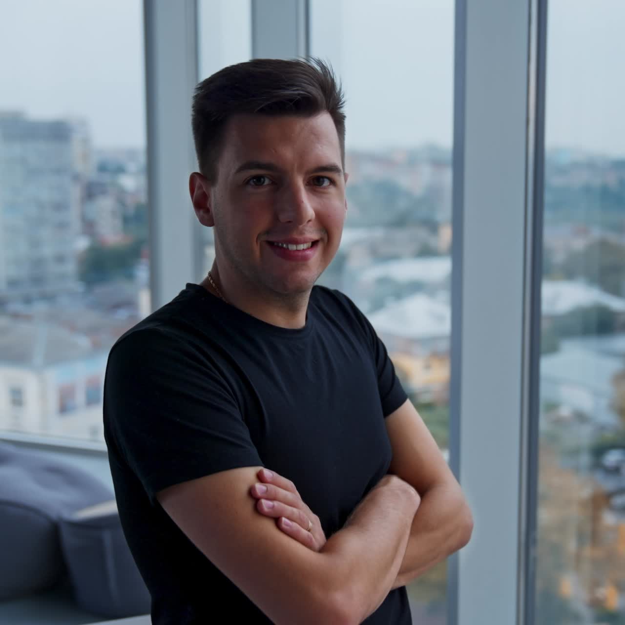 Approaching the young man sitting on the desk in office. Male looking into window and then turns to the camera smiling. Blurred backdrop