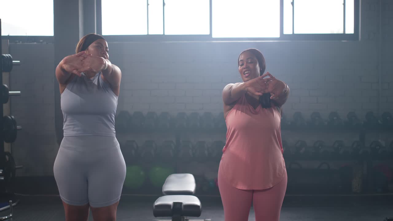 Amiche con top rosa e blu che eseguono un riscaldamento per le braccia in palestra vicino ai manubri prima dell'allenamento.