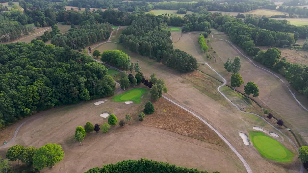 Aerial drone orbits over Bramshaw Golf Course in Brook, Hampshire, revealing greens, dry brown fairways, trees, and houses in the distance. Ideal for golf, sports, or drought-themes
