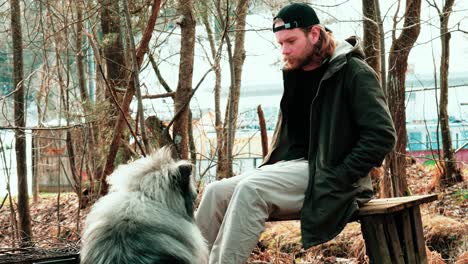 a-blond-young-man-in-a-cap-lights-a-cigarette-on-a-bench-while-his-fluffy-friend-watches-him