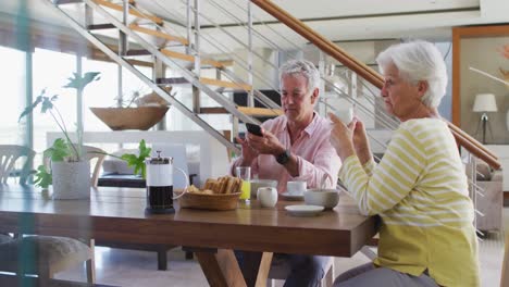 Pareja-Caucásica-Mayor-Hablando-Entre-Sí-Usando-Un-Teléfono-Inteligente-Desayunando-Juntos-En-Casa