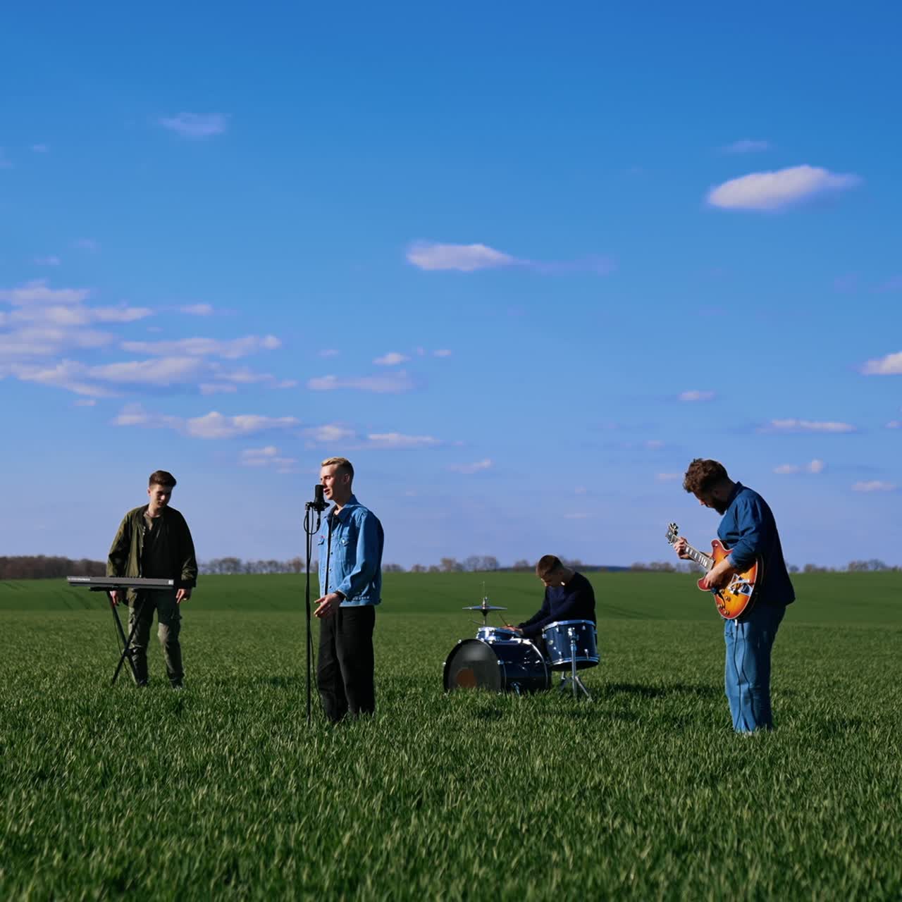Outdoor concert of young musicians. Group of friends playing the guitar, drums and synthesizer performing music on green field under blue sky