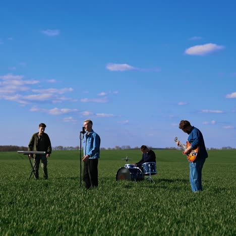 outdoor concert of young musicians. group of friends playing the guitar, drums and synthesizer performing music on green field under blue sky