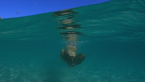 Half-underwater-scene-of-little-girl-with-diving-mask-swimming-in-clear-turquoise-seawater-toward-camera-splashing-sea-water