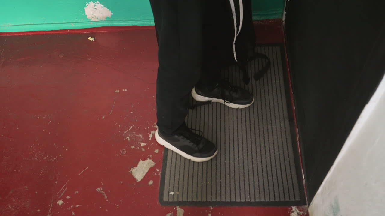 Sorte sneakers træder på dørmåtte ved døråbningen, rødt slidt gulv og afskallet grøn vægramme gummimåtte, nærbillede i overliggende engel fanger kurér-ankomstsekvens, tom måtte efterfulgt af fodplacering, kort