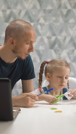 pretty little girl cuts out paper figures while father looks at daughter sitting at white table with modern laptop in brightly lit room staying home
