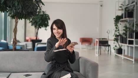 young,-beautiful-woman-in-a-suit-is-sitting-on-the-sofa-in-the-business-center's-lobby,-happily-and-joyfully-scattering-dollar-bills