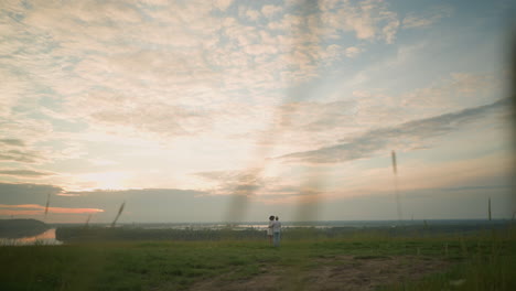 a couple stands on a grassy hill beside a lake at sunset, gazing at the sky. the man, in a white shirt, hat, and jeans, gently holds the woman by the waist. she wears a black hat and a white dress