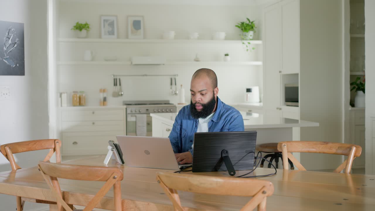 African American man blue-shirted sitting in kitchen responding to email on laptop and monitor